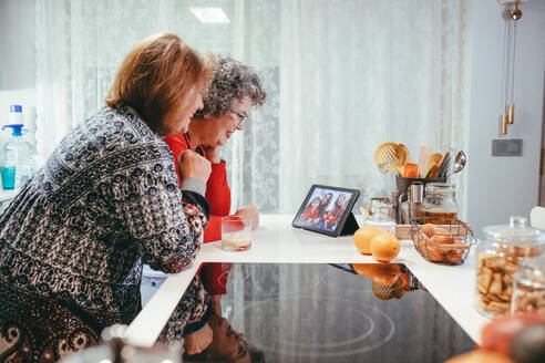 Side view of smiling homosexual elderly women watching tablet with happy children and mothers during video chat at home - ADSF23330