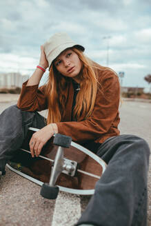 Young female skater with long blond hair in trendy outfit sitting on asphalt road with cruiser skateboard in hand against cloudy sky in countryside - ADSF23336