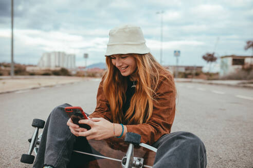 Glücklicher weiblicher Teenager mit langen roten Haaren in trendigem Outfit und Hut, der auf einem Skateboard sitzt und lächelt, während er ein Selfie mit seinem Handy macht - ADSF23338