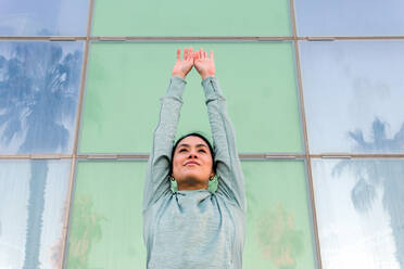 Selbstbewusste junge ethnische Sportlerin in modischer Sportkleidung, die wegschaut, während sie vor einem modernen Glasgebäude auf einer Stadtstraße steht - ADSF23343