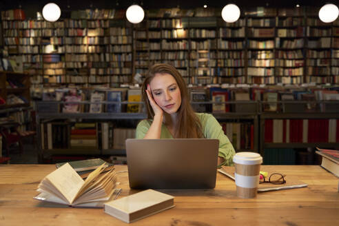 Woman with head in hand looking at laptop in library - VEGF04363