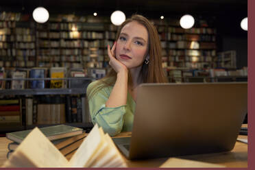 Schöne Frau mit Hand am Kinn in der Bibliothek sitzend - VEGF04366