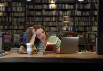 Frau mit Hand im Haar, die in einer Bibliothek ein Buch liest - VEGF04368