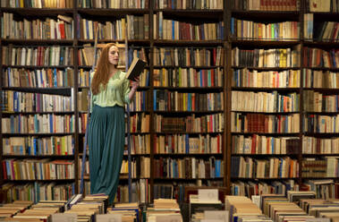 Engagierte Frau liest ein Buch, während sie auf einer Leiter in einer Bibliothek steht - VEGF04387