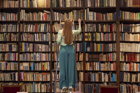 Young woman standing on ladder while searching book in library - VEGF04390