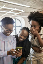 Glückliche weibliche Fachkraft, die ein Mobiltelefon benutzt, während ihre Kolleginnen im Coworking-Büro lachen - ZEDF04145