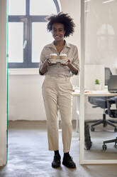 Afro-Geschäftsfrau, die Einweg-Kaffeebecher in der Hand hält, während sie am Eingang eines Büros steht - ZEDF04191
