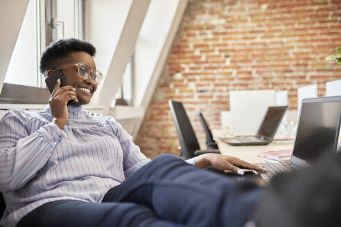 Weibliche Fachkraft mit Brille, die mit einem Smartphone spricht, während sie einen Laptop im Büro benutzt - ZEDF04214