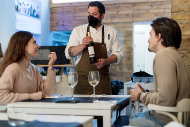 Ehepaar im Gespräch mit dem Kellner, der eine Weinflasche im Restaurant hält, während COVID-19 - EGAF02334