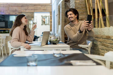 Lächelnder Mann, der ein Selfie mit seinem Smartphone macht, während er seine Freundin im Restaurant ansieht - EGAF02353