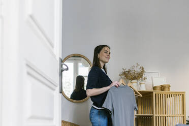 Smiling woman contemplating while holding clothes in vacation home - XLGF01599