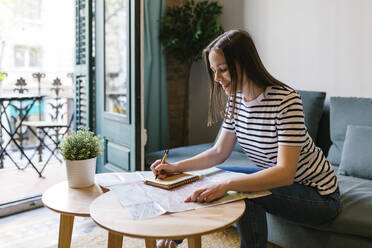 Lächelnde Frau schreibt eine Notiz in ihr Tagebuch, während sie am Tisch in einer Wohnung sitzt - XLGF01605