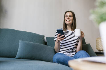 Smiling woman having coffee while holding mobile phone on sofa in vacation home - XLGF01606
