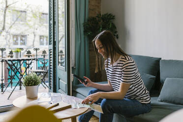 Smiling female tourist holding mobile phone while reading newspaper in vacation apartment - XLGF01608