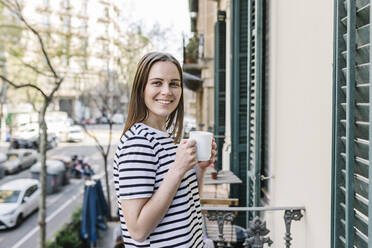 Happy woman having coffee while standing in balcony - XLGF01610