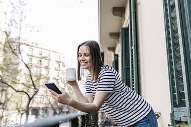 Lächelnde Frau mit Kaffeetasse, die ein Mobiltelefon benutzt, während sie sich auf einem Balkon an ein Geländer lehnt - XLGF01612