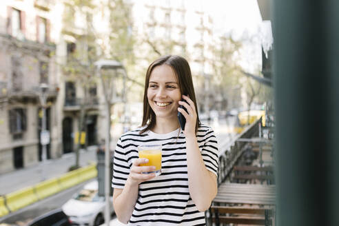 Lächelnde junge Frau, die einen Saft trinkt, während sie auf einem Balkon mit ihrem Handy telefoniert - XLGF01618