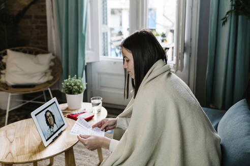 Kranke Frau, eingewickelt in eine Decke, spricht während einer telemedizinischen Behandlung über ein digitales Tablet zu Hause - XLGF01623