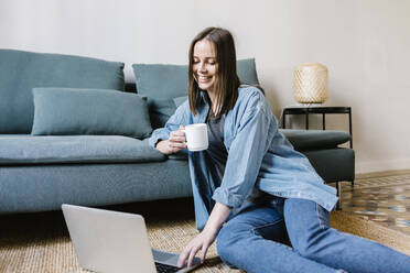 Lächelnde junge Frau, die einen Laptop benutzt, während sie zu Hause auf dem Sofa Kaffee trinkt - XLGF01638