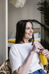 Woman day dreaming while holding bottle of juice in living room - XLGF01641