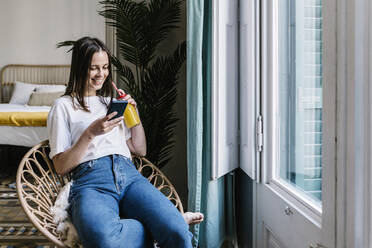 Young woman having juice while using smart phone on chair at home - XLGF01643