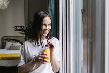 Smiling young woman looking away while having orange juice by window at home - XLGF01645