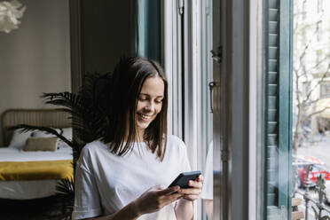 Smiling young woman using smart phone by window at home - XLGF01648