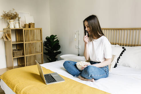 Woman eating popcorn while looking at laptop on bed at home - XLGF01651