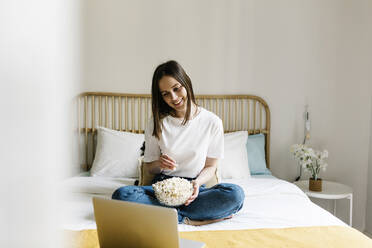 Lächelnde junge Frau, die Popcorn isst und auf einen Laptop auf dem Bett schaut - XLGF01653