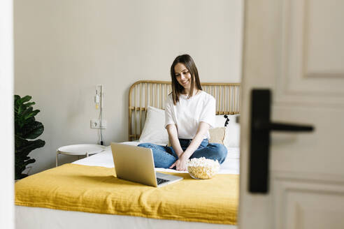 Junge Frau schaut auf einen Laptop, während sie zu Hause auf dem Bett sitzt - XLGF01654