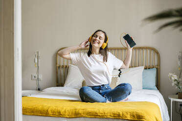 Cheerful woman with mobile phone listening music through headphones in bedroom - XLGF01666