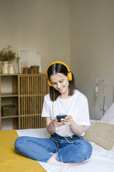 Woman with cross legs using smart phone while listening music through headphones in bedroom - XLGF01667
