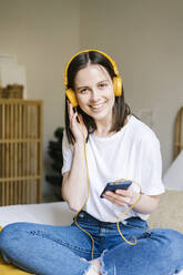 Happy woman listening music through headphones while holding mobile phone on bed at home - XLGF01668