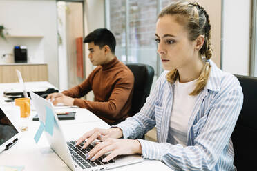 Junge Geschäftsfrau, die einen Laptop benutzt, während sie mit einem Kollegen im Hintergrund im Büro sitzt - XLGF01671