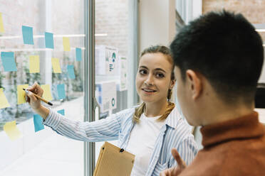 Lächelnde Unternehmerinnen und Unternehmer, die über Haftnotizen an der Glaswand eines Coworking-Büros diskutieren - XLGF01682