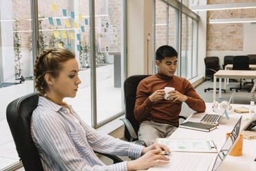 Businesswoman working on laptop by male coworker having coffee in office - XLGF01696