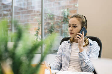 Young entrepreneur talking on mobile phone while working at office - XLGF01698