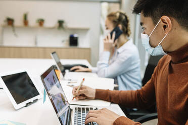 Businessman wearing protective face mask working on laptop with female colleague in background at office - XLGF01703