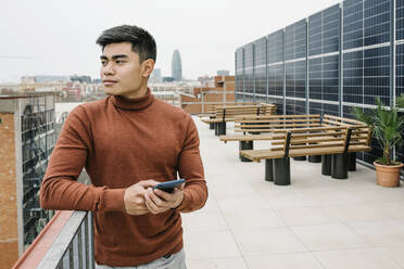 Male entrepreneur holding mobile phone while standing at rooftop - XLGF01729