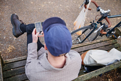 Mann benutzt sein Smartphone auf dem Fahrrad, während er auf einer Bank sitzt - ASGF00244