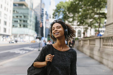 Lächelnde Frau, die wegschaut, während sie auf einem Fußweg in der Stadt über Kopfhörer Musik hört - BOYF01977
