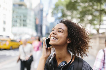 Lächelnde Frau mit Kopfhörern, die in der Stadt mit einem Smartphone telefoniert - BOYF01980