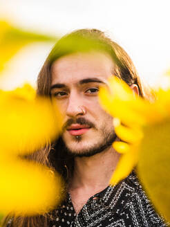 Serene unemotional hipster male with long hair standing in yellow sunflower field and looking at camera - ADSF23347