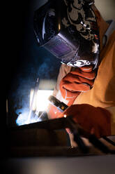 Unbekannter männlicher Angestellter mit Schutzhandschuhen und Helm bei der Arbeit an einem Schweißgerät in einer dunklen Werkstatt - ADSF23372