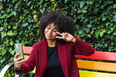 Inhalt Afroamerikanische Frau sitzt auf einer Bank in der Stadt und nimmt ein Selbstporträt mit ihrem Smartphone auf, während sie die Geste der zwei Finger zeigt - ADSF23373