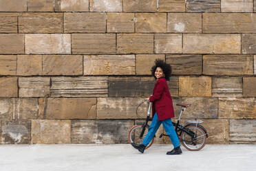 Side view of positive black female walking with bicycle on street along old stone wall and looking at camera - ADSF23375