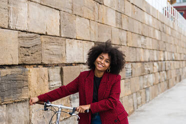 Front view of positive black female walking with bicycle on street along old stone wall and looking at camera - ADSF23376