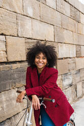 Vorderansicht einer positiven schwarzen Frau, die mit einem Fahrrad auf der Straße entlang einer alten Steinmauer geht und in die Kamera schaut - ADSF23377