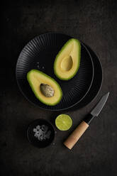 Frische Avocadohälften von oben auf einem Tisch mit grüner Limette, Salz und Messer auf dunklem Hintergrund angeordnet - ADSF23404