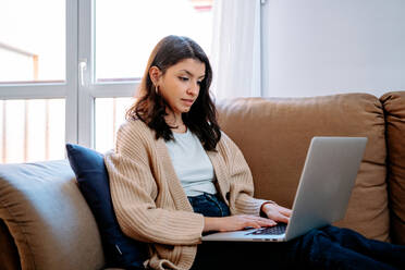 Side view of focused female freelancer sitting on couch at home and working on remote project while typing on laptop - ADSF23413